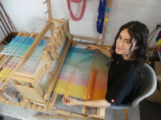Une jeune femme souriante, passionnée par son tissier métier, travaille sur un métier à tisser en bois. Les fils colorés créent une œuvre vibrante.