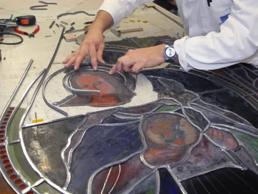 Un artisan assemble un vitrail, joignant des pièces de verre coloré avec du plomb. Le visage d'une Vierge et d'un enfant se dessine.