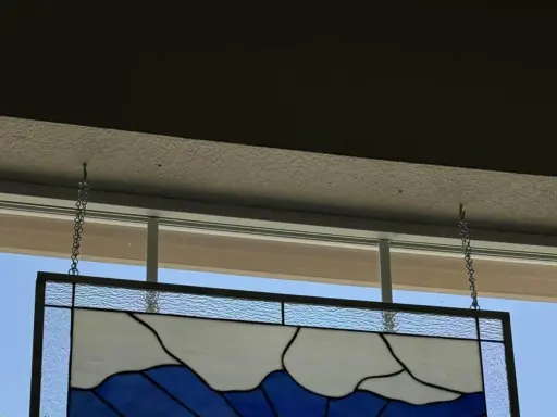 Comment fixer un vitrail sur une fenêtre : un panneau de verre bleu et blanc est suspendu par des chaînes à des crochets fixés au plafond.