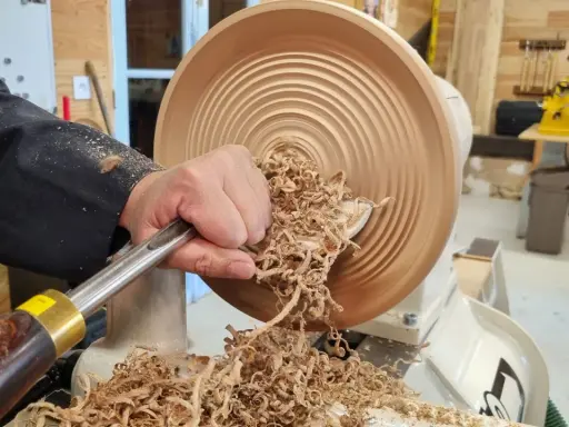 Main d'un artisan façonnant un objet en bois sur un tour. Les copeaux volent, témoignant du travail de tournage sur bois.