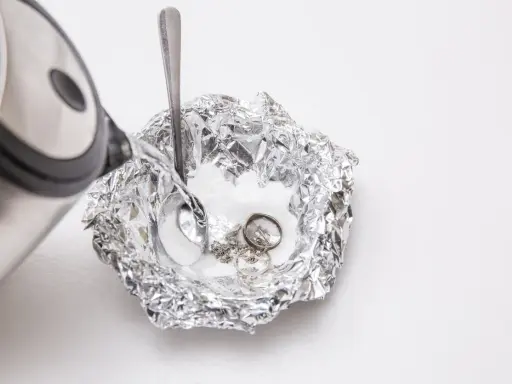 Nettoyage d'argenterie noircie : de l'eau chaude est versée sur des bijoux en argent et une cuillère dans une coupelle en papier aluminium.