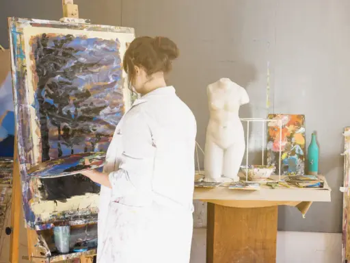 Un artisan artiste, en blouse blanche, tient une palette devant une toile abstraite dans son atelier. Une statue de femme est visible.