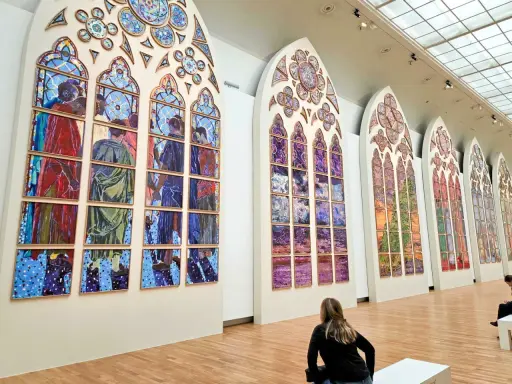 Des vitraux contemporains de Notre-Dame, des œuvres vibrantes composées de multiples panneaux peints, évoquant des scènes humaines et des paysages colorés.