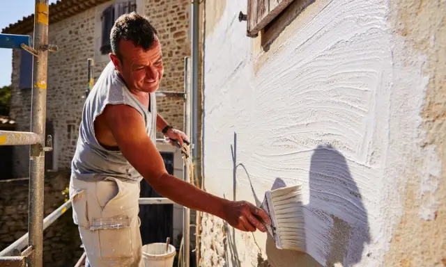 Un homme applique un badigeon blanc sur un mur en pierre avec un large pinceau. Le soleil éclaire la scène, créant des ombres.