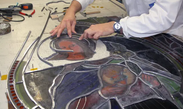 Un artisan assemble un vitrail, joignant des pièces de verre coloré avec du plomb. Le visage d'une Vierge et d'un enfant se dessine.