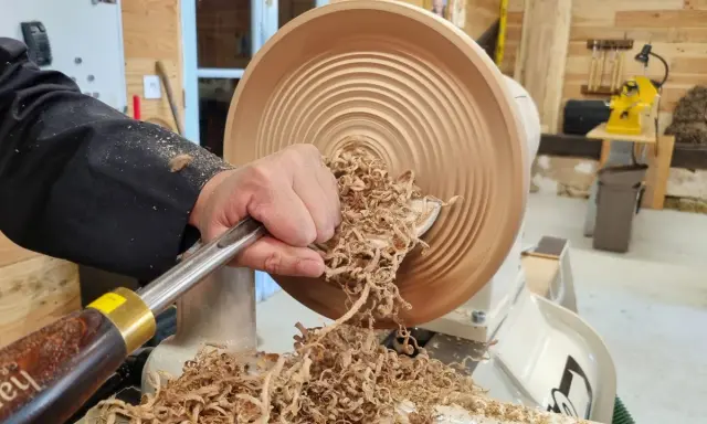 Main d'un artisan façonnant un objet en bois sur un tour. Les copeaux volent, témoignant du travail de tournage sur bois.