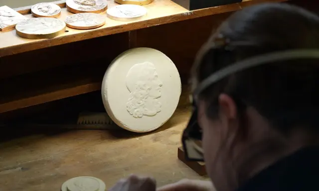 Un graveur métier travaille sur un moule en plâtre orné d'un profil antique. D'autres médailles sont visibles sur l'établi.