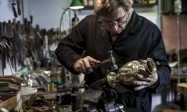 Un orfèvre au travail, martelant délicatement un vase en métal dans son atelier rempli d'outils.