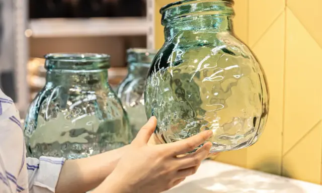 Une main examine un objet en verre texturé, vert pâle, aux reflets ondulés. D'autres objets similaires sont visibles en arrière-plan.