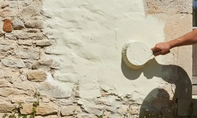 Une main applique une peinture claire sur un mur en pierre enduit de chaux. La question "peut on peindre sur de la chaux" trouve ici sa réponse visuelle.
