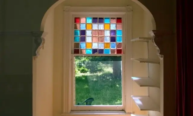Un vitrail design aux carrés colorés illumine une fenêtre, projetant des reflets joyeux sur l'herbe verdoyante à l'extérieur.