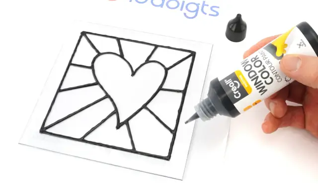 Main noire pour un modèle vitrail en forme de cœur. Un tube de peinture contour est tenu par une main.