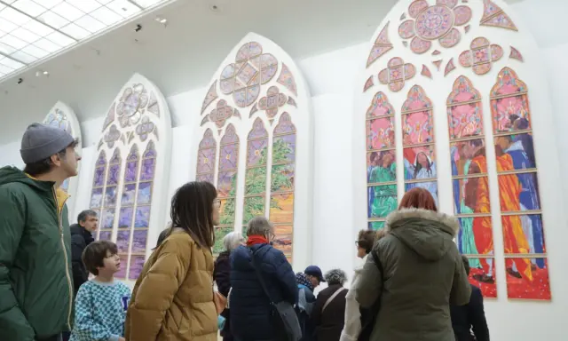 Des visiteurs contemplent de grands vitraux contemporains aux couleurs vives, représentant des scènes et des paysages.