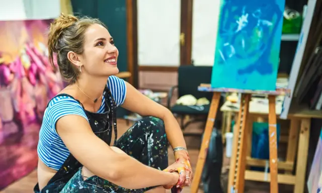 Une artiste rêveuse, les mains tachées de peinture, s'interroge sur comment devenir artisan d'art. Son atelier est rempli de toiles colorées.