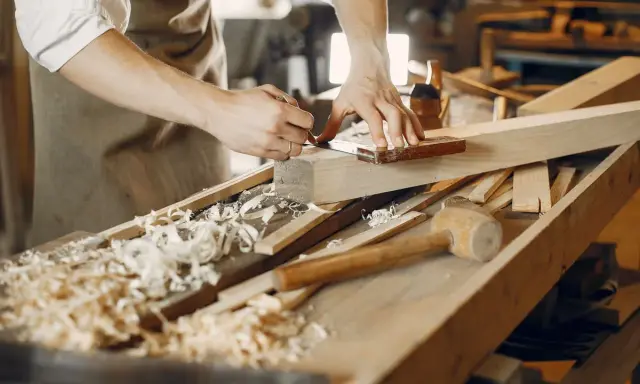 Un artisan qui travaille le bois, concentré sur la précision de ses gestes. Des copeaux de bois jonchent l'établi.