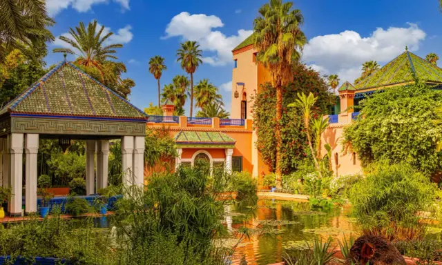 Jardin luxuriant avec un étang reflétant une maison bleue à Marrakech, palmiers et ciel bleu.