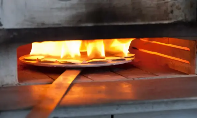 Des pièces de métal attendent d'être émaillées dans un four, sous des flammes vives. L'artisan émailleur surveille attentivement la cuisson.
