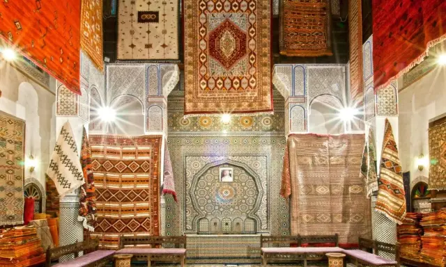 Un intérieur richement décoré de tapis colorés, témoignant de l'artisanat de Fès. Des motifs géométriques et floraux ornent les murs et les sols.