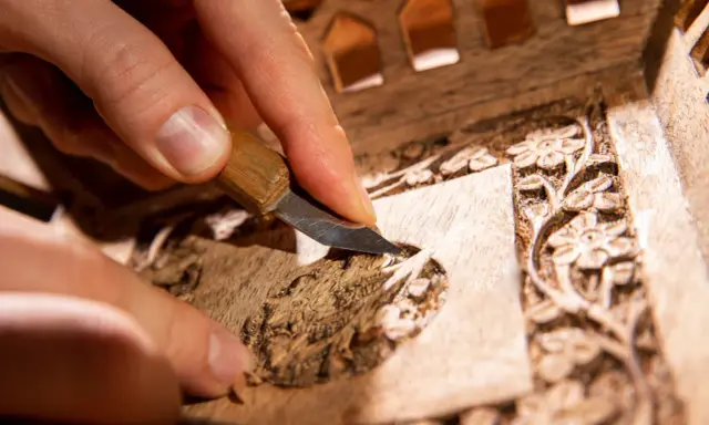 Main d'artisan sculptant un détail floral sur un meuble en bois, étape clé pour savoir comment nettoyer un meuble en marqueterie.