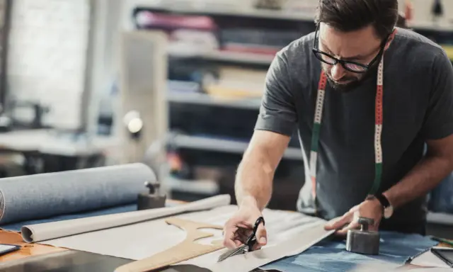 Un artiste textile concentré, lunettes sur le nez, coupe un tissu avec précision dans son atelier.