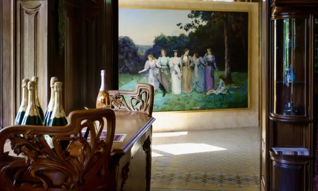 Bouteilles de champagne sur une table en bois sculpté, devant une peinture de femmes dans un jardin, ambiance Villa Mademoiselle Reims.