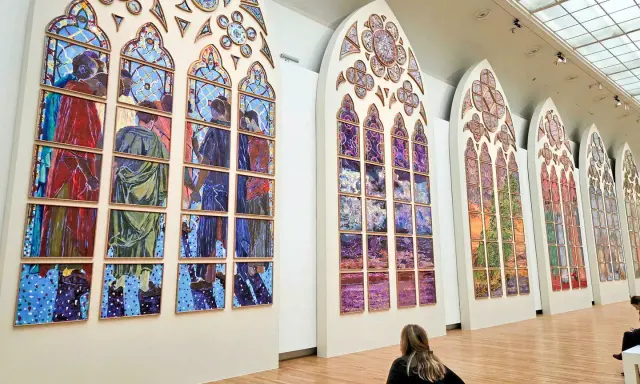 Des vitraux contemporains de Notre-Dame, des œuvres vibrantes composées de multiples panneaux peints, évoquant des scènes humaines et des paysages colorés.