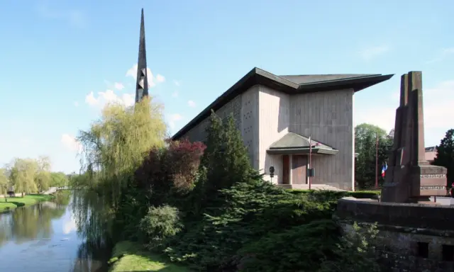 L'église Saint-Rémy de Baccarat, avec sa flèche élancée, se dresse près d'une rivière bordée d'arbres verdoyants.