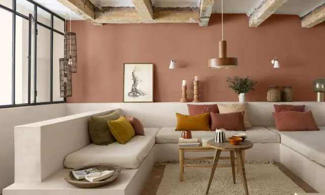 Salon cosy aux murs couleur brun terre de France, banquette en L, coussins ocre et rouille, table basse en bois brut.