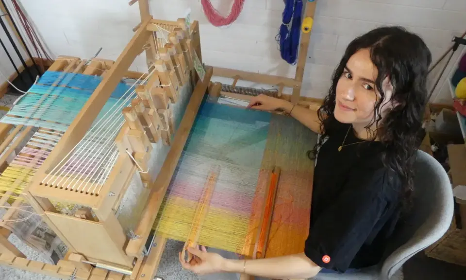 Une jeune femme souriante, passionnée par son tissier métier, travaille sur un métier à tisser en bois. Les fils colorés créent une œuvre vibrante.