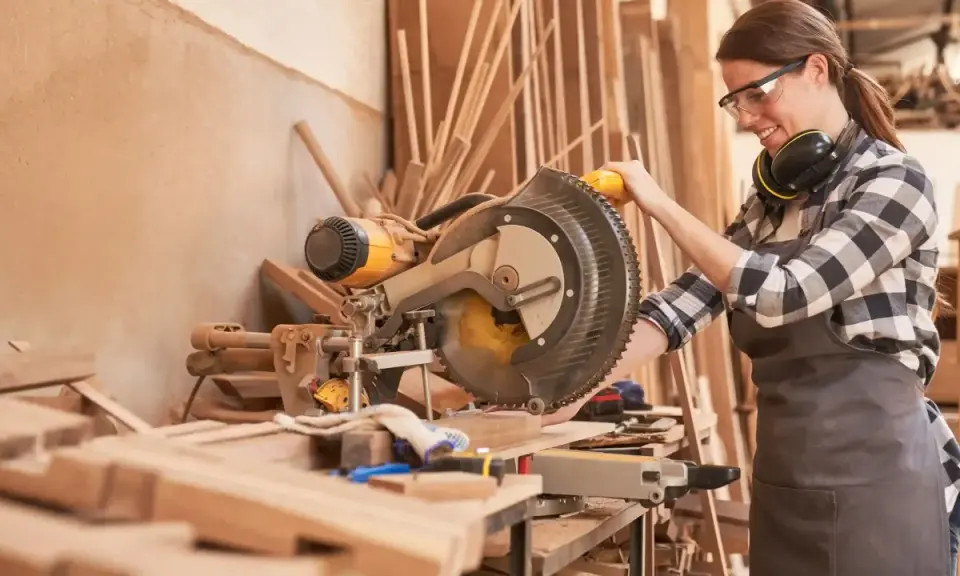 Artisane souriante utilisant une scie à onglet. Elle porte des lunettes et un tablier, des éléments essentiels pour comment protéger une création artisanale.