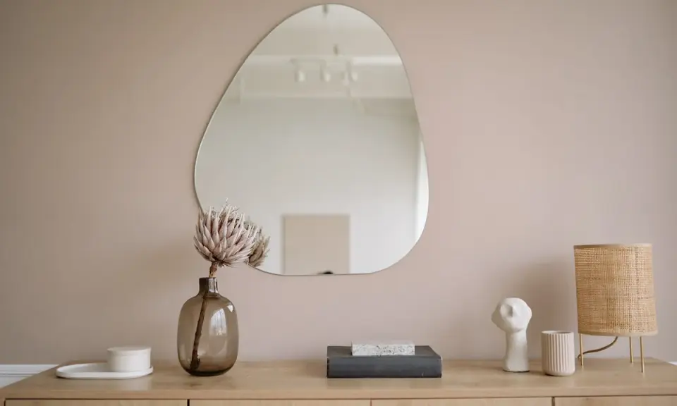 Un miroir vitrail aux formes organiques reflète une pièce épurée. Une fleur séchée dans un vase en verre fumé et une lampe en rotin complètent la scène.