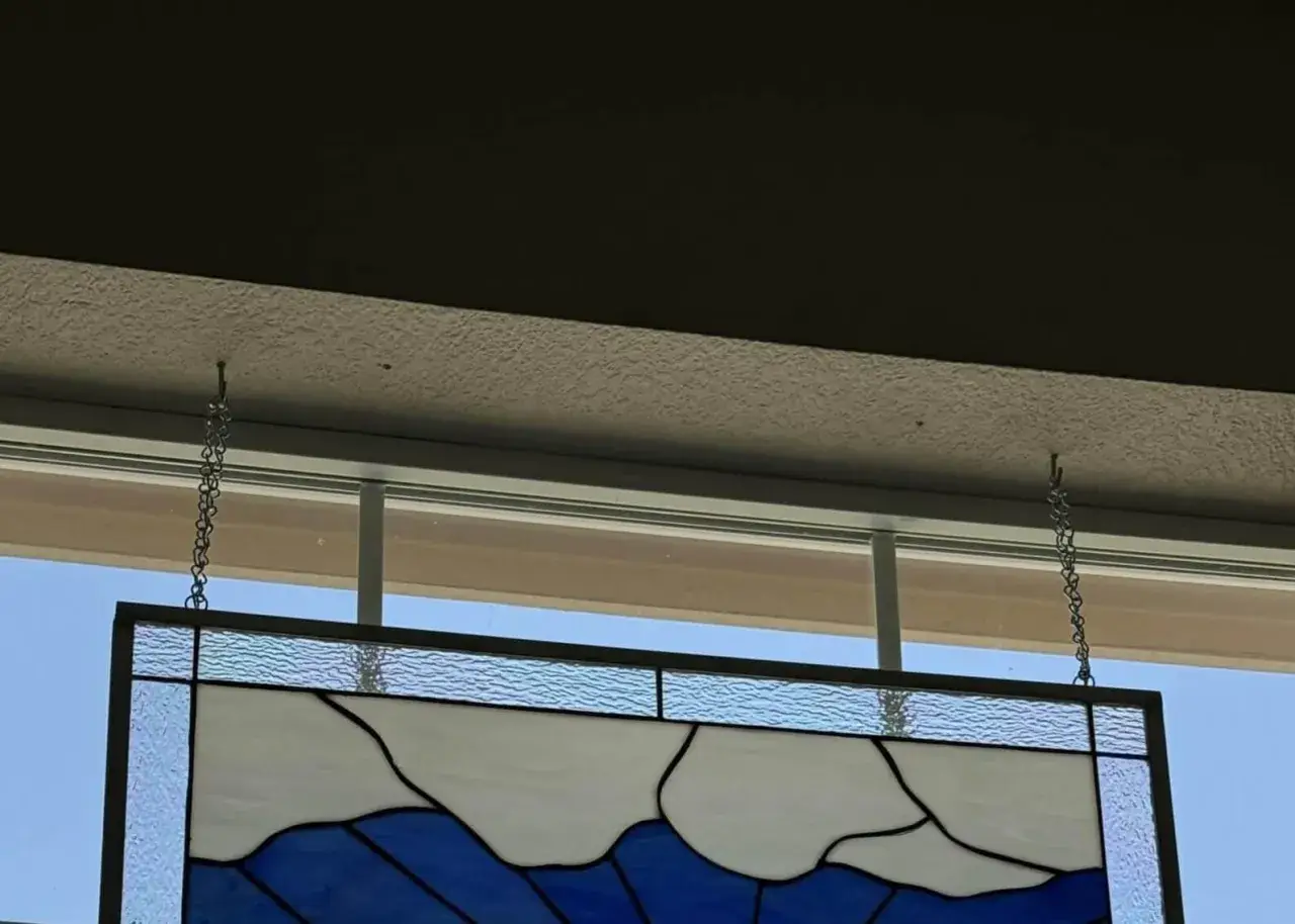 Comment fixer un vitrail sur une fenêtre : un panneau de verre bleu et blanc est suspendu par des chaînes à des crochets fixés au plafond.