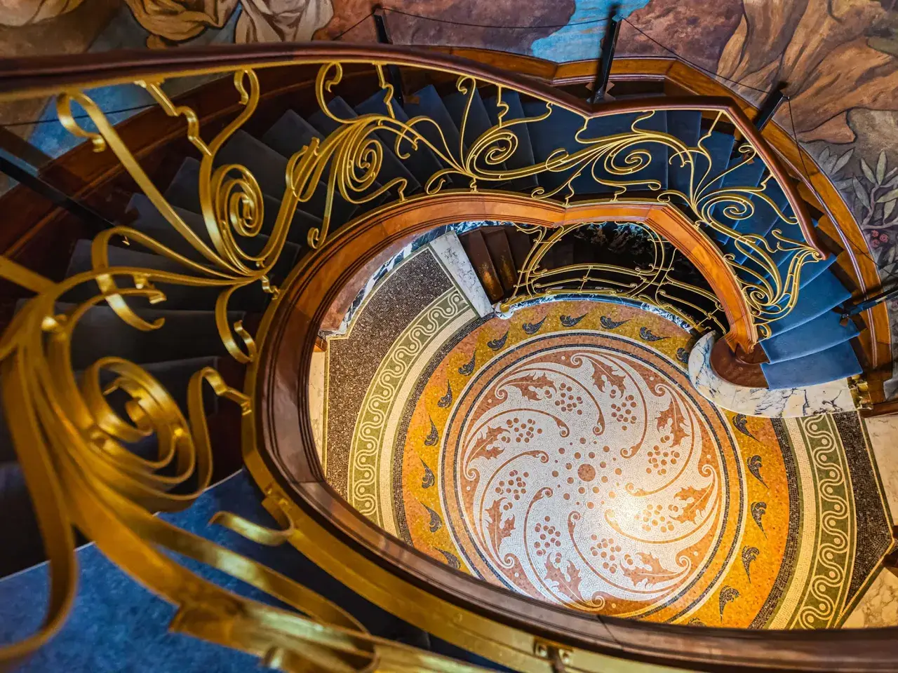 Escalier en colimaçon orné d'une rampe dorée aux volutes végétales, rappelant une fleur art nouveau. Le sol en mosaïque circulaire est un chef-d'œuvre.