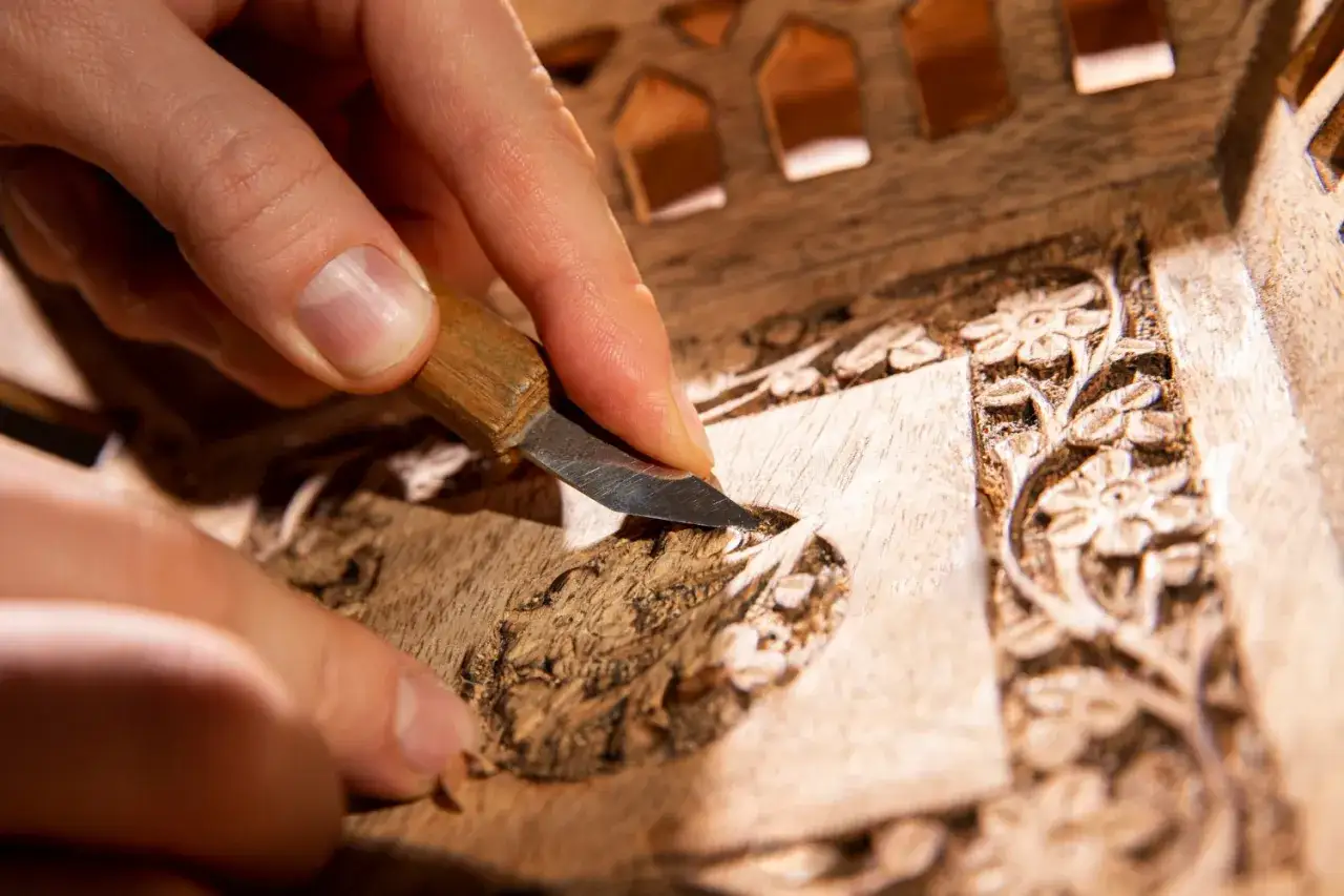 Main d'artisan sculptant un détail floral sur un meuble en bois, étape clé pour savoir comment nettoyer un meuble en marqueterie.