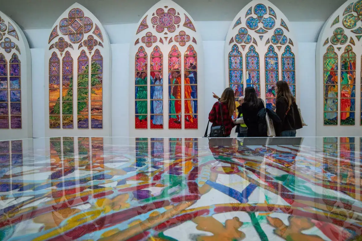 Trois personnes admirent des vitraux contemporains aux couleurs vives, leurs reflets se mêlant à une œuvre au sol.