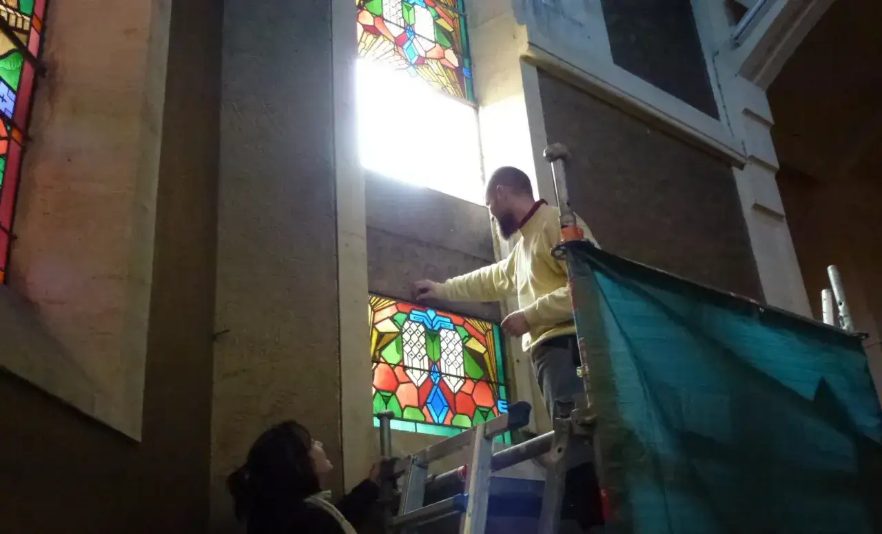 Un homme sur un &eacute;chafaudage ajuste un vitrail color&eacute; dans une &eacute;glise. Une femme regarde.