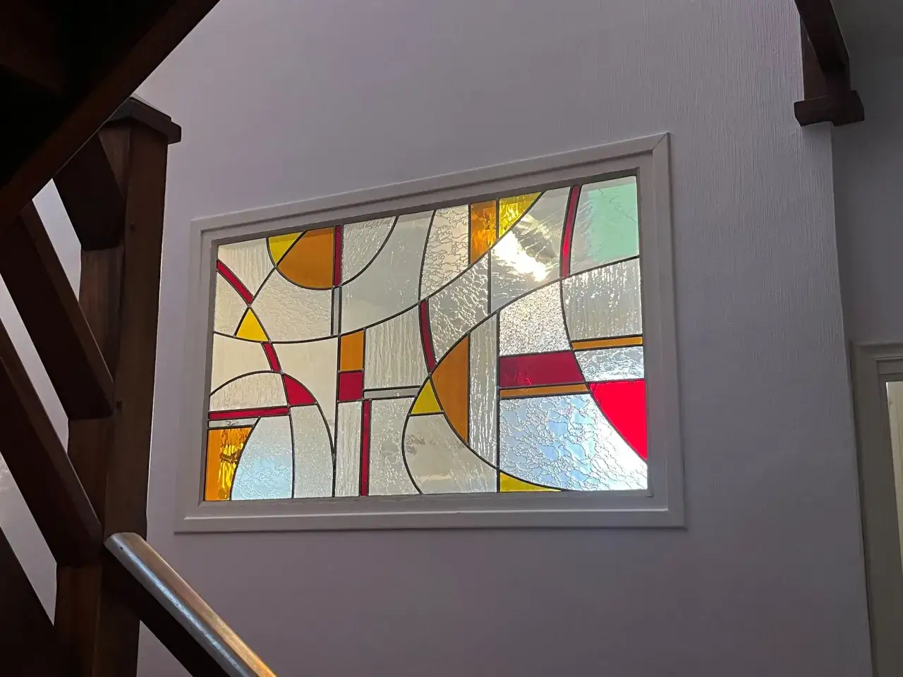 Un vitrail abstrait aux couleurs vives éclaire un escalier en bois. Le dessin vitraux joue avec les formes géométriques et les textures.