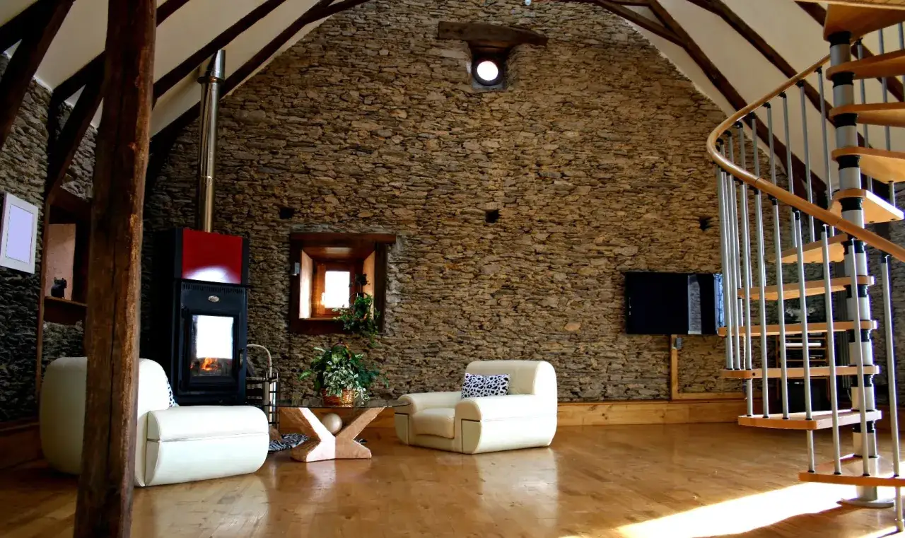 Salon rustique avec mur en pierre naturelle, poêle, fauteuils blancs et escalier en colimaçon. Le nettoyage pierre naturelle intérieur est essentiel pour préserver ce charme.