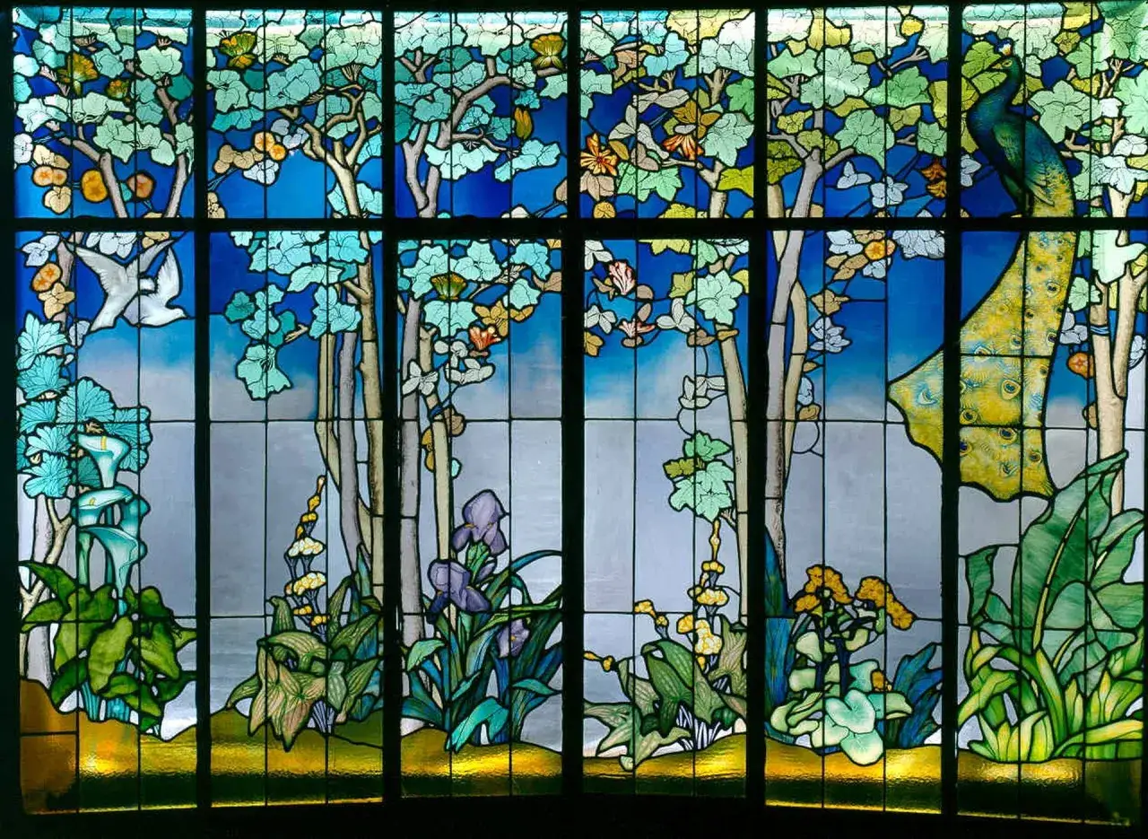 Un magnifique vitrail design Art Nouveau dépeint une scène naturelle avec un paon, des arbres, des fleurs et un oiseau blanc.