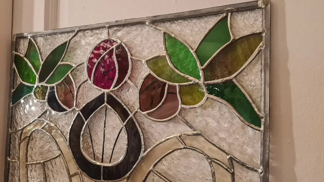 Un vitrail aux fleurs stylis&eacute;es, aux teintes vertes, pourpres et brunes, capture la lumi&egrave;re.