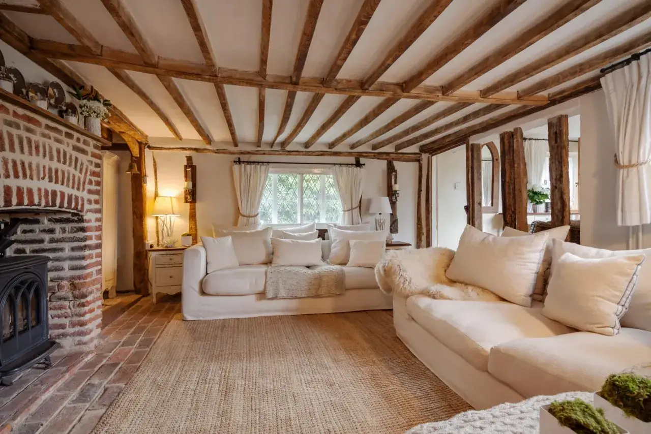 Salon douillet style cottage avec chemin&eacute;e en brique, poutres apparentes et canap&eacute;s blancs confortables.
