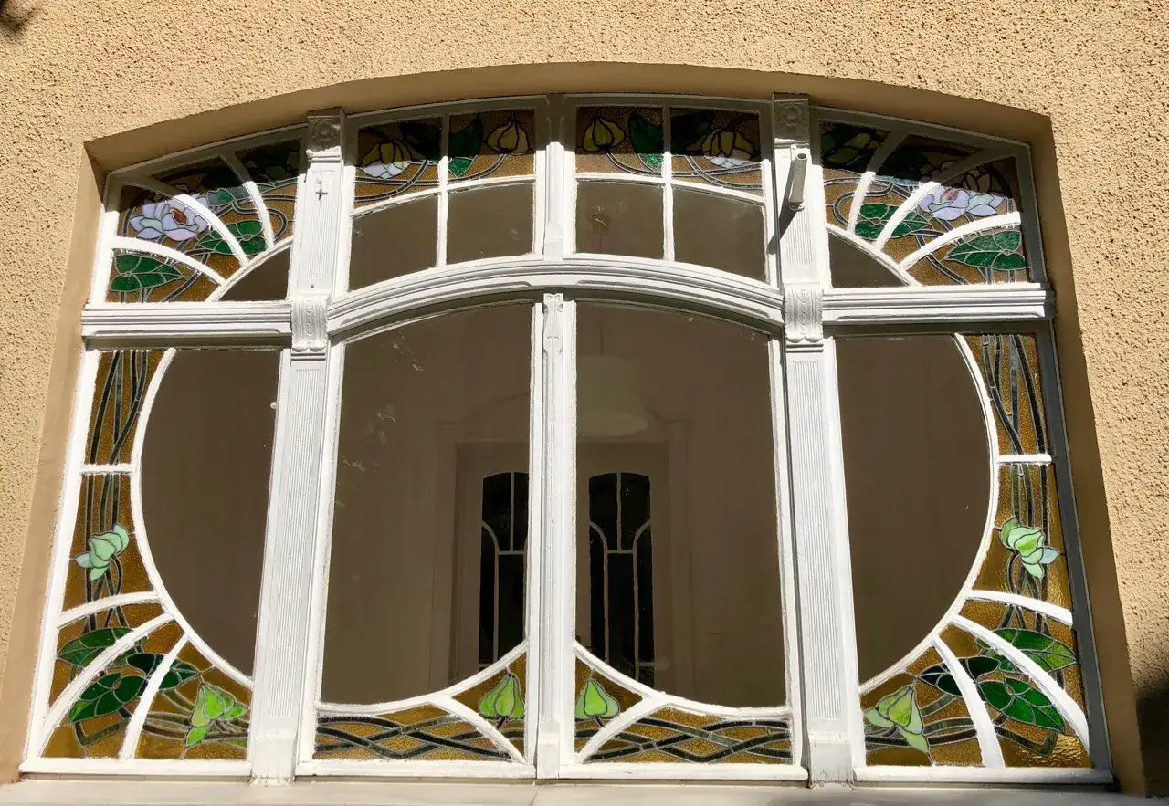 Magnifique fenêtre Art Nouveau ornée de vitraux aux motifs floraux et de lignes courbes, encadrée de blanc sur un mur texturé.