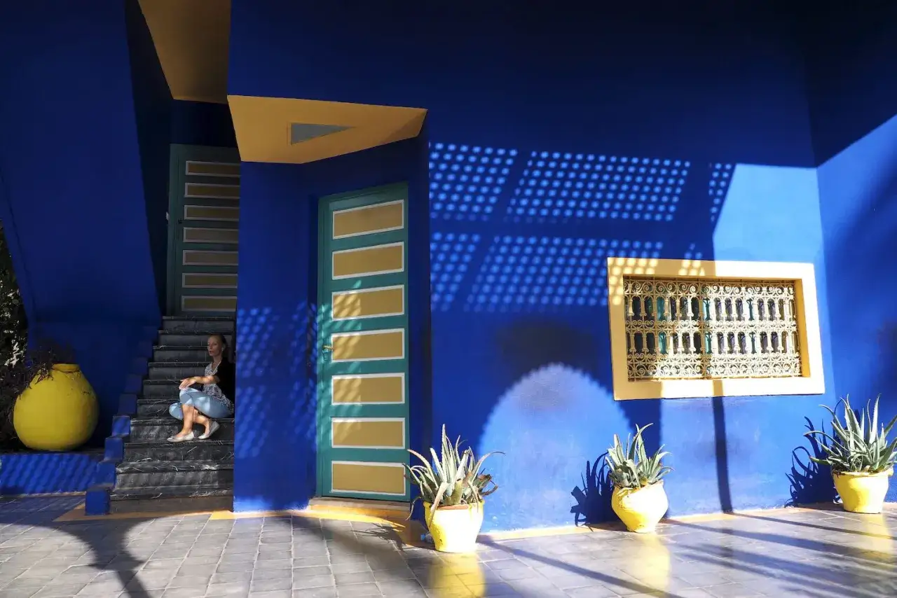 Une femme assise sur les marches d'une maison bleue &agrave; Marrakech, avec des ombres d&eacute;coratives projet&eacute;es sur le mur.