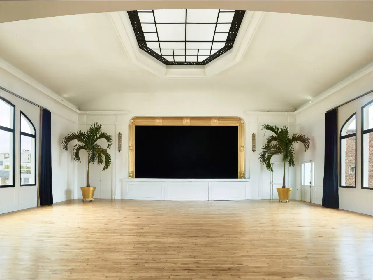 Vaste salle avec parquet, deux palmiers en pots dor&eacute;s, et une sc&egrave;ne orn&eacute;e d'une entr&eacute;e art d&eacute;co.