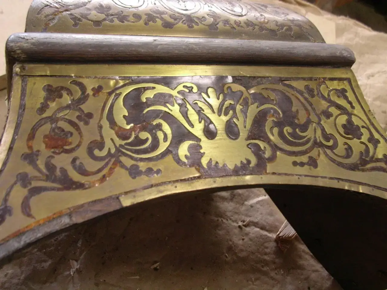Détail d'un meuble ancien en bois sombre avec incrustations de laiton aux motifs floraux. On observe des traces de patine et des zones où comment décaper du laiton serait nécessaire pour révéler son éclat.