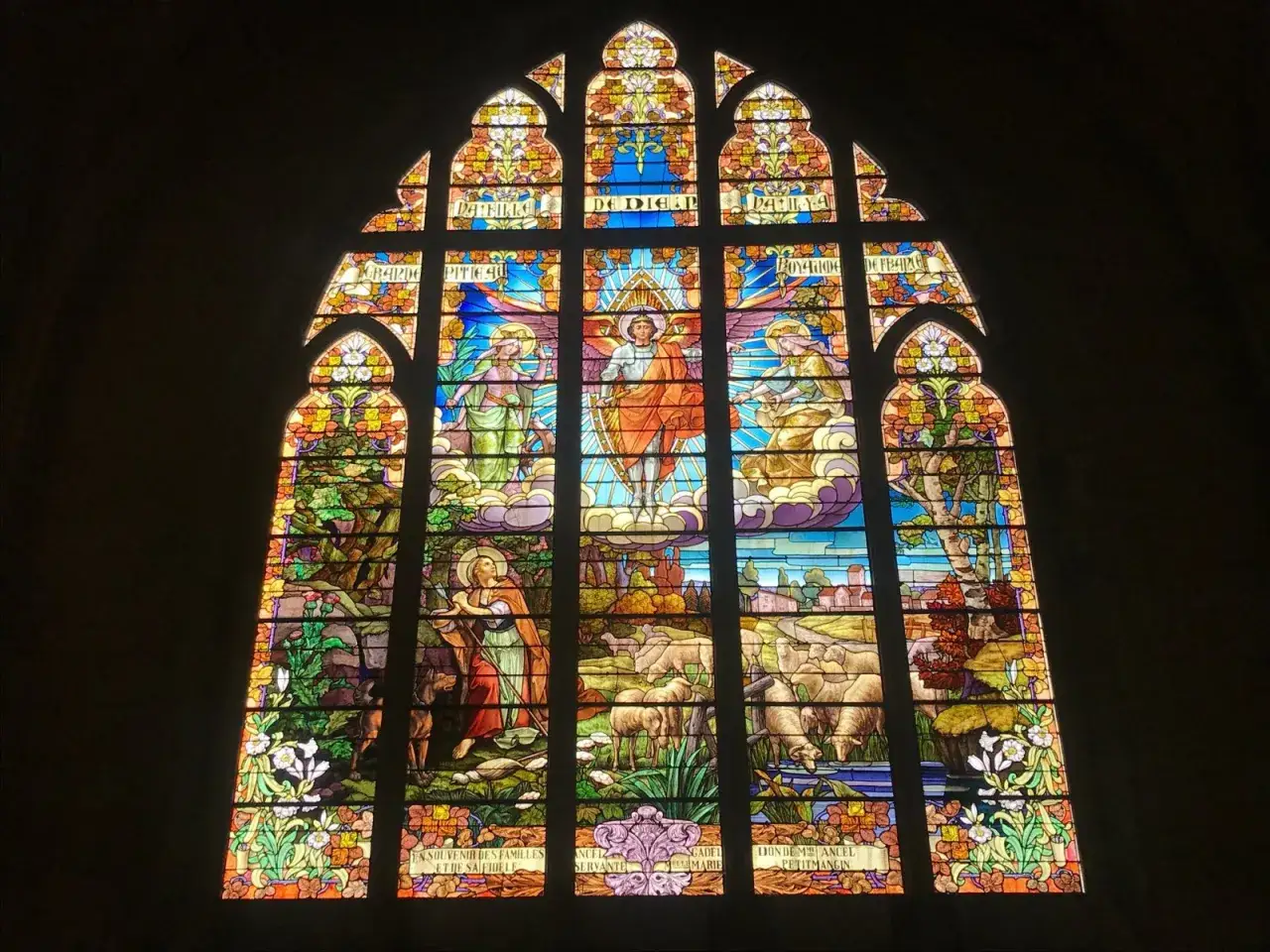 Vitrail de Jeanne d'Arc : une sc&egrave;ne c&eacute;leste avec des anges et une figure centrale en armure, entour&eacute;e de moutons et d'un paysage bucolique.
