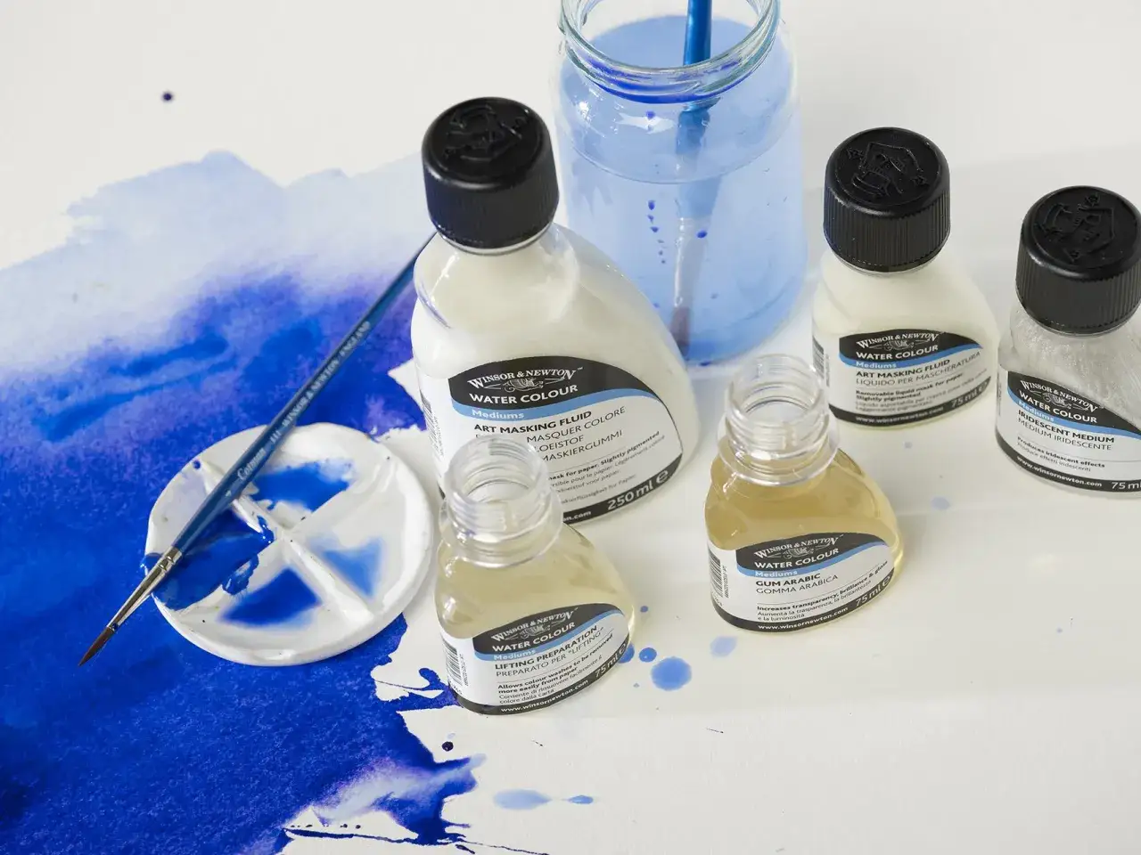 Pinceau sur palette avec des peintures bleues. Bouteilles de m&eacute;diums pour aquarelle, dont la colle gomme arabique, sur fond blanc.
