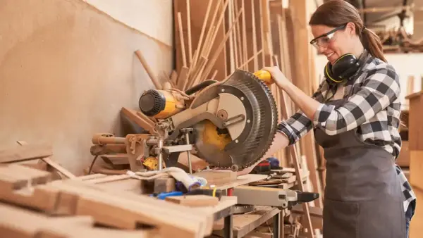 Artisane souriante utilisant une scie à onglet. Elle porte des lunettes et un tablier, des éléments essentiels pour comment protéger une création artisanale.
