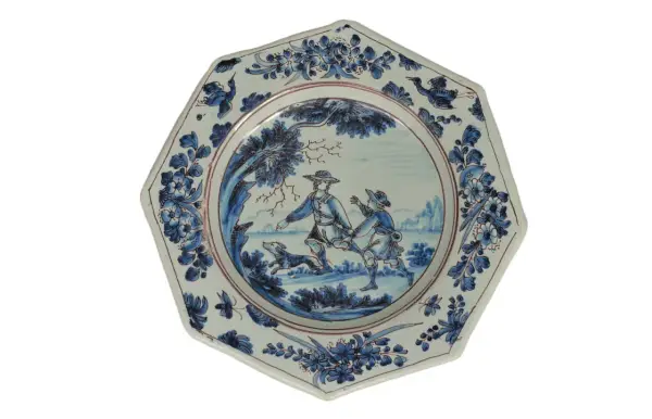 Assiette octogonale en céramique de Marseille, décorée de deux hommes et d'un chien dans un paysage champêtre, bordée de motifs floraux bleus.