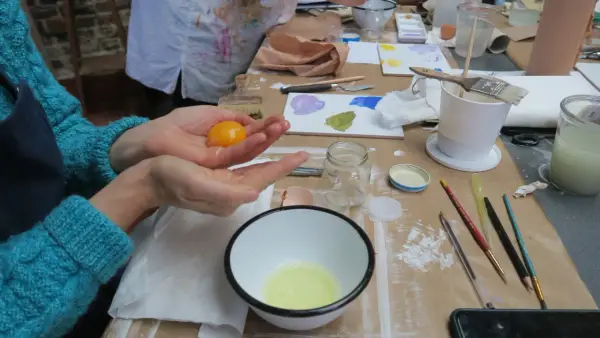Préparation d'une tempera peinture à l'oeuf : un jaune d'œuf est tenu dans une main au-dessus d'un bol contenant le blanc.